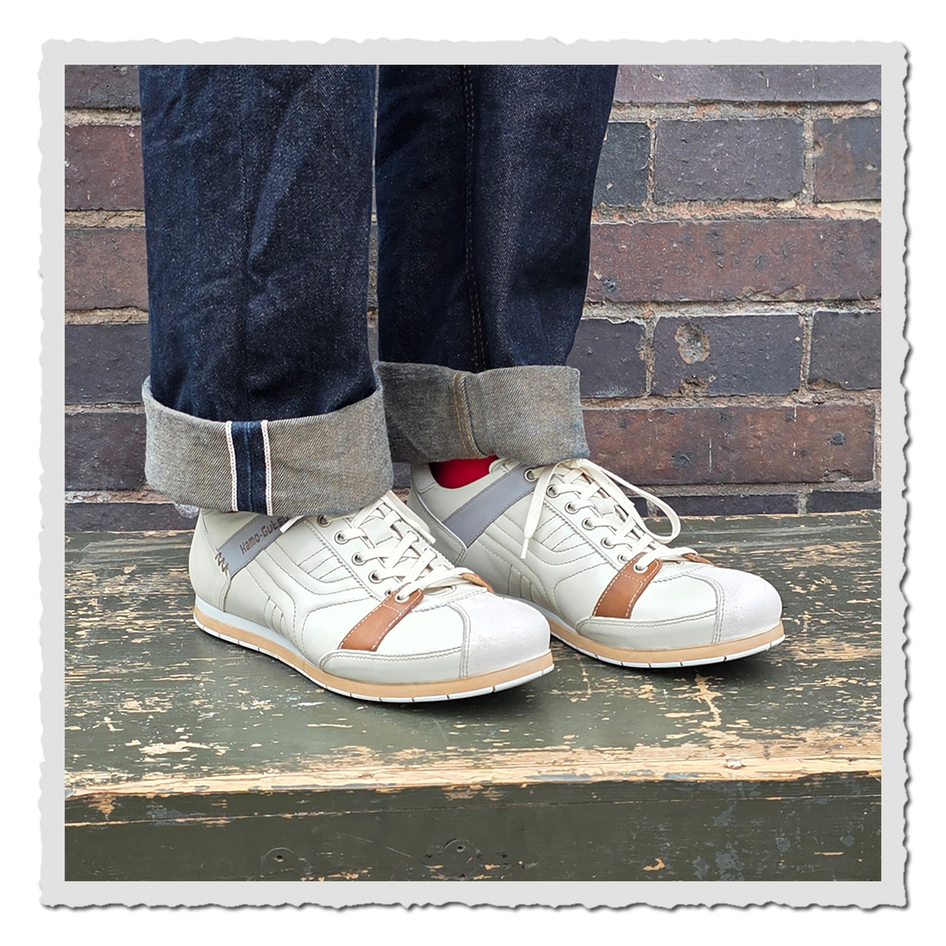 Altweiße Schnürschuhe an einer Person mit umgeschlagenen dunklen Jeans, auf grün lackierter Holzfläche vor Backsteinwand.